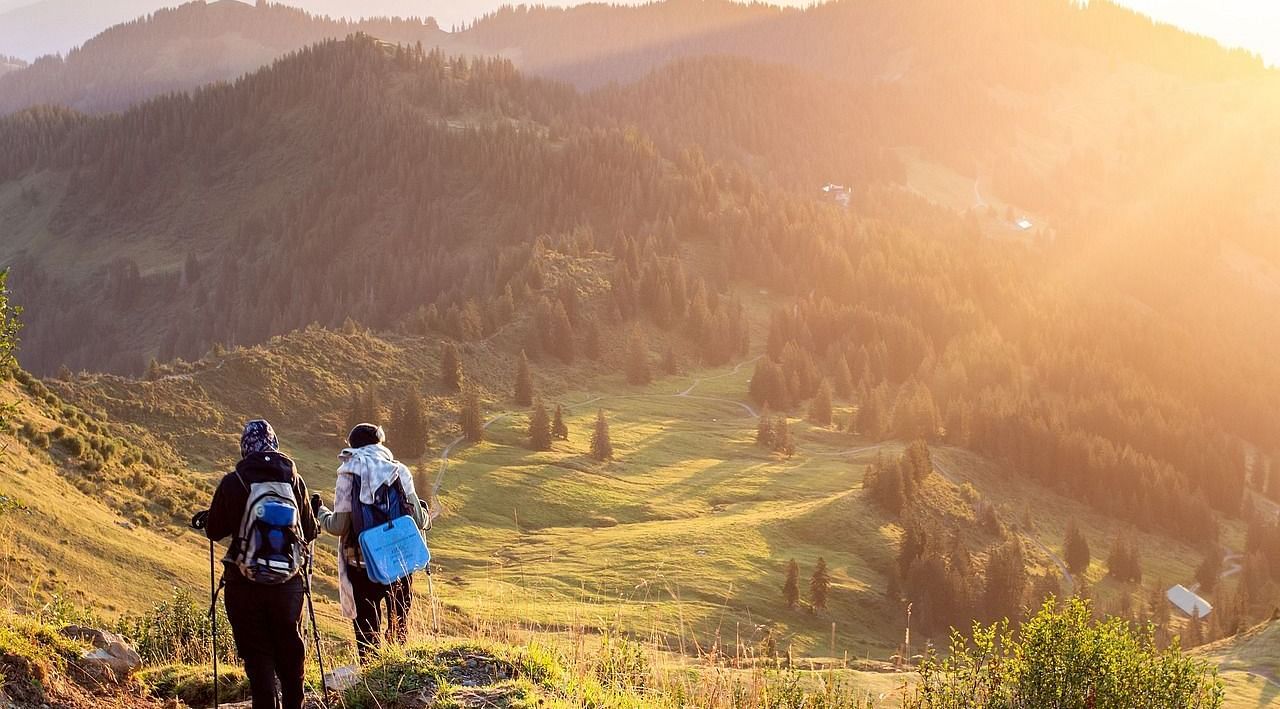 Two hikers walk down a sunlit mountain trail, surrounded by rolling hills and forests, bathed in the warm glow of sunset.