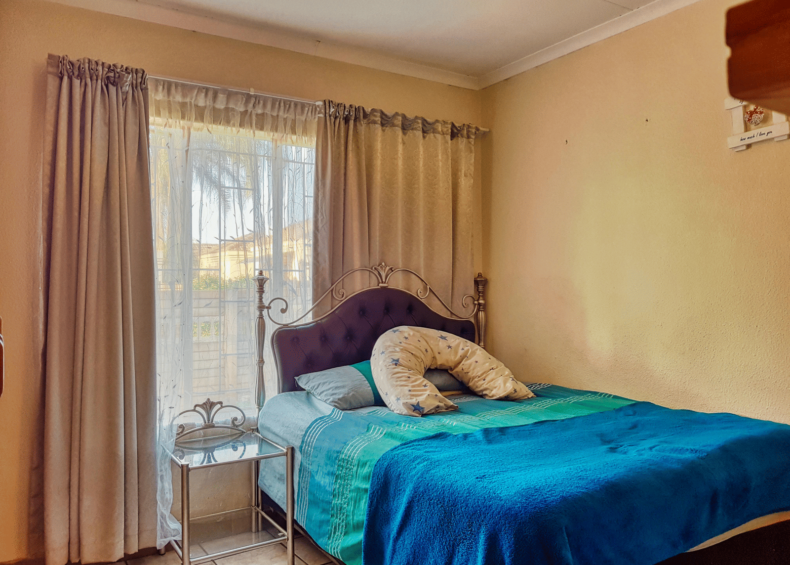 A bedroom featuring a double bed with blue sheets spread neatly over it with beige curtains covering the window behind it.