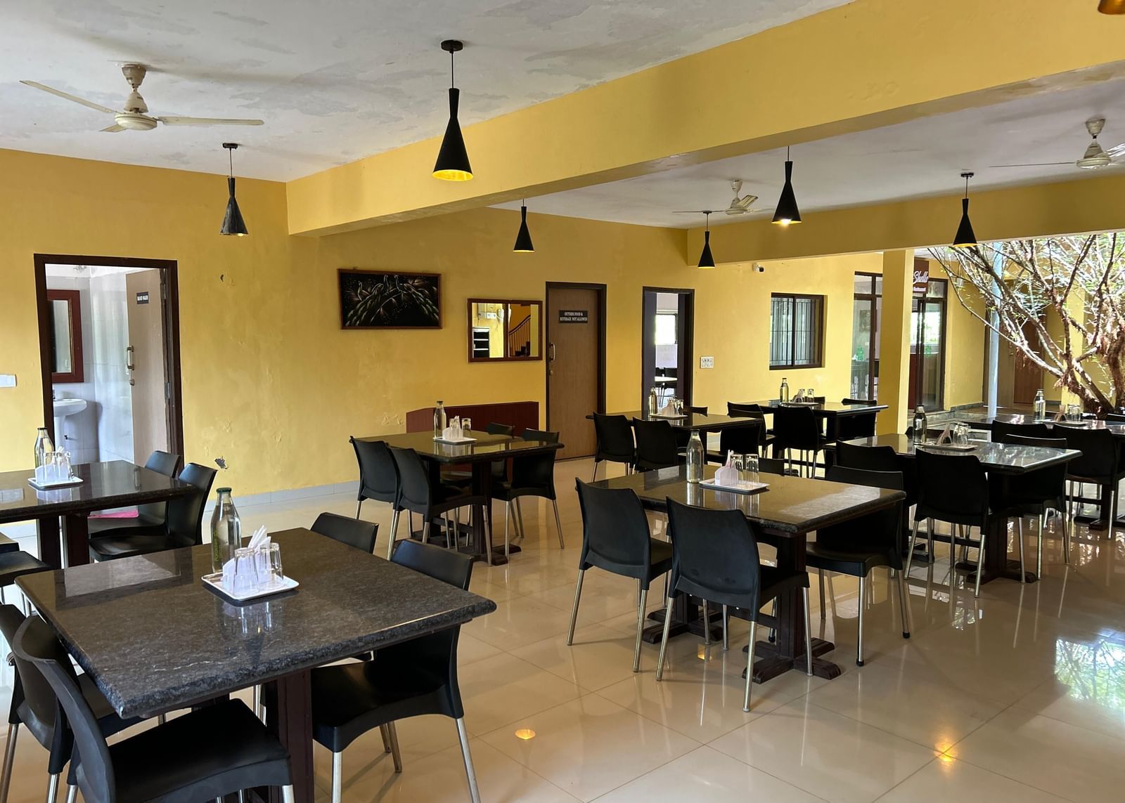 Dining room with yellow walls, tables, black chairs, and hanging lights at Paradise Wild Hills Resort, Kollur.
