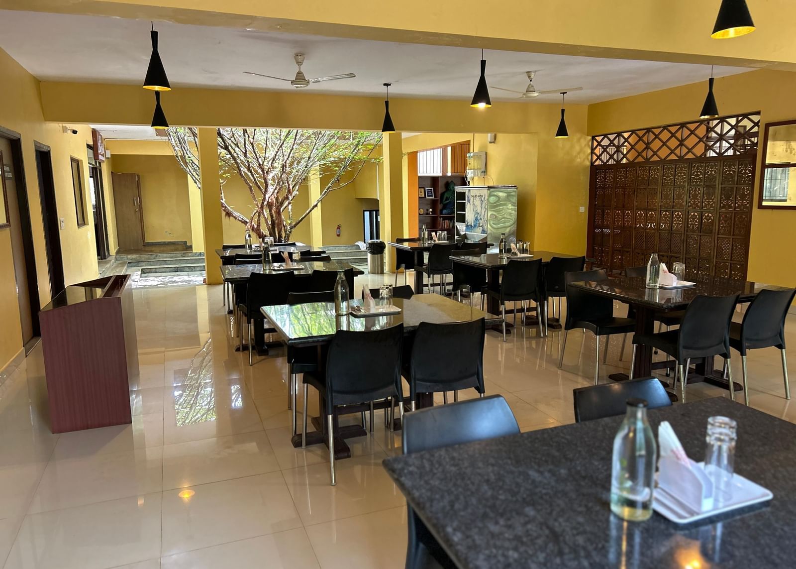 Dining area with yellow walls, tables, chairs, and indoor tree feature at Paradise Wild Hills Resort, Kollur.
