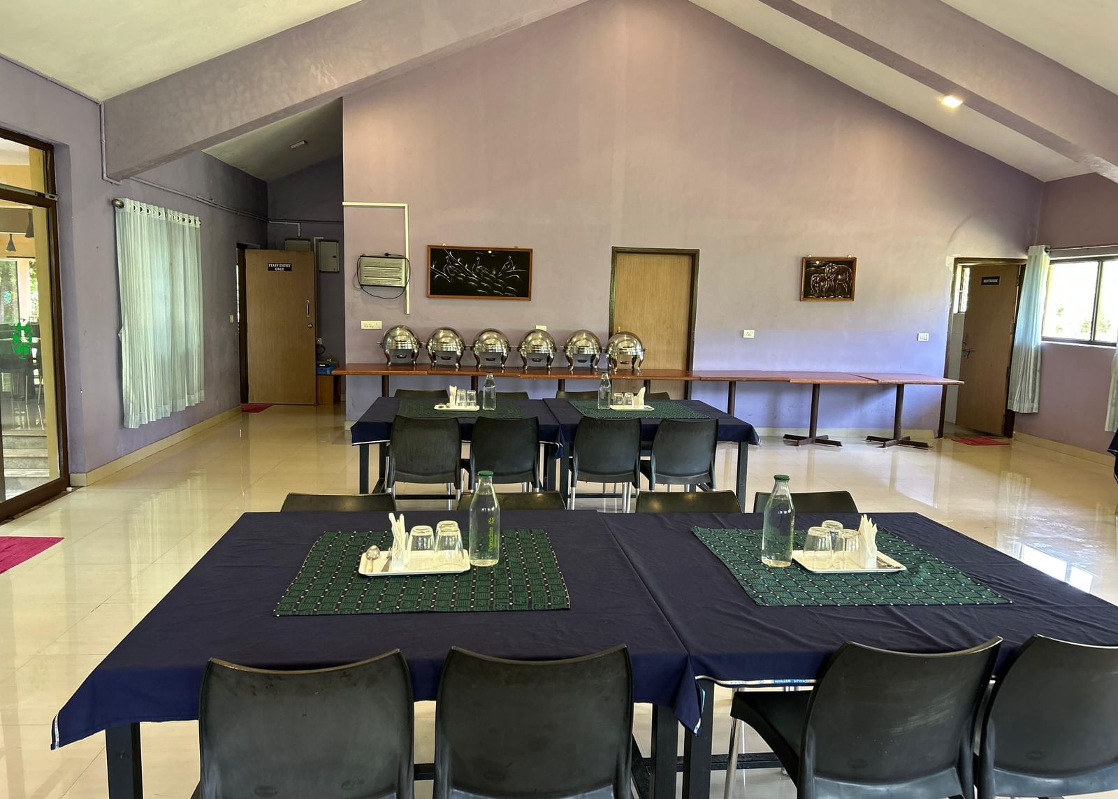 Indoor dining space with light purple walls, covered tables, chairs, and wide windows at Paradise Wild Hills Resort, Kollur.