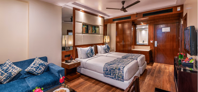 A hotel room is furnished with two single beds, a sofa, a table with snacks, a television, and wooden flooring. 