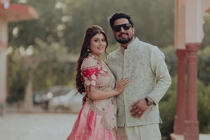 A couple poses elegantly in traditional attire, sharing a warm smile in an outdoor setting with a serene background. - Umaid Palace - A Lakeside Heritage Palace Resort