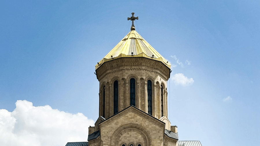 A large, multi-tiered stone cathedral featuring a golden dome and cross set against a bright, slightly cloudy blue sky.
