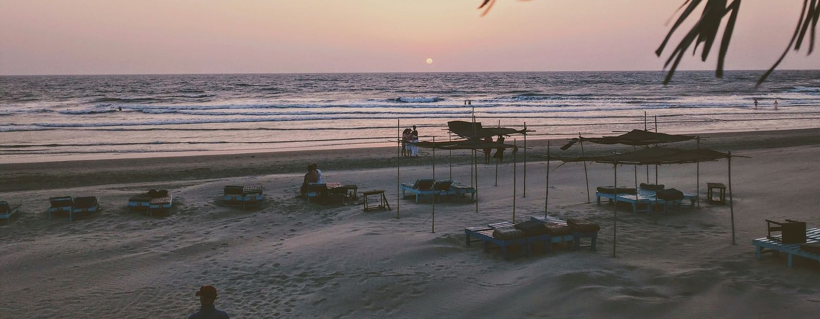 A tranquil beach scene at sunset, with lounge chairs and huts silhouetted against the fading light over the ocean.