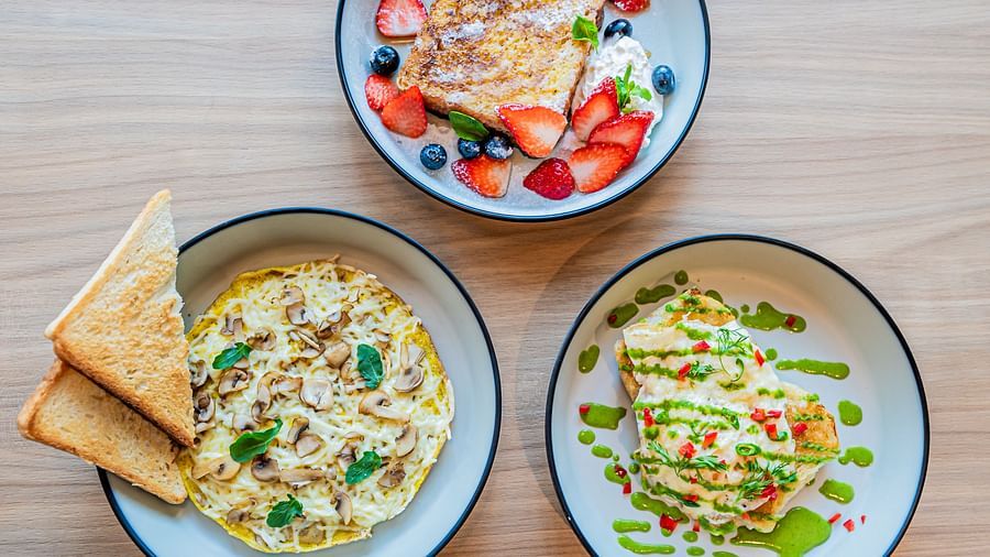 Three plates at Indie Stays Mumbai International Airport: one with mushroom omelette and basil leaves, one with toast topped with strawberries and blueberries, and one with egg toast and veggies.