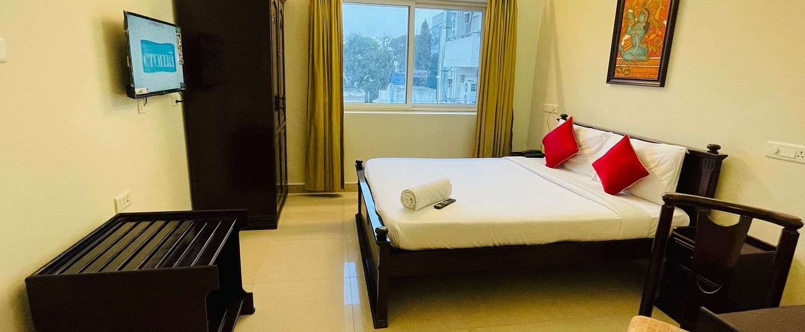 A well-lit room with a bed dressed in white linens and accented with red cushions, alongside wooden furniture and a wall-mounted television at Sanctum Suites Indiranagar, Bangalore.