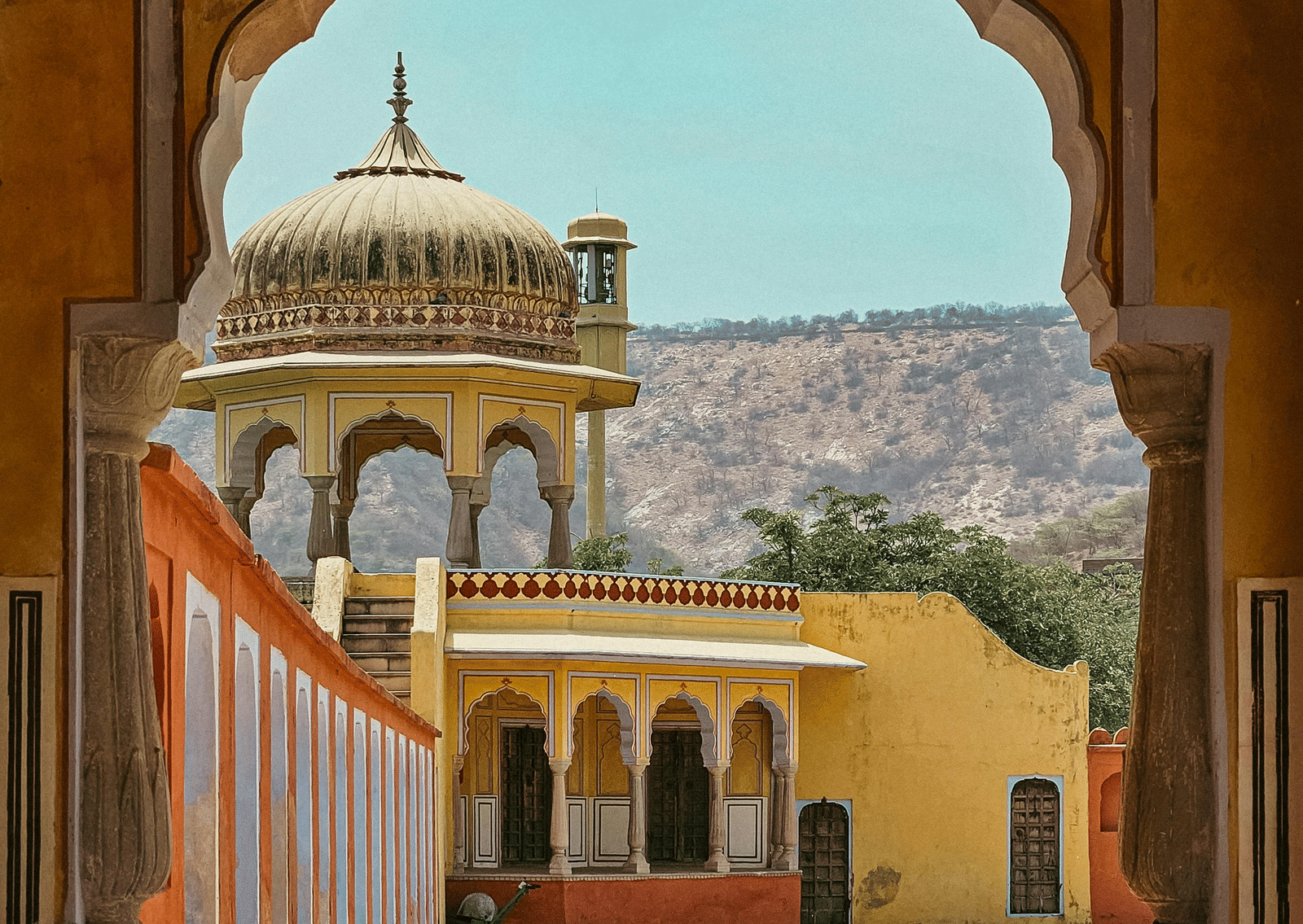 An ornate, arched yellow and orange sandstone doorway leading out onto a balcony, overlooking hills in the background.