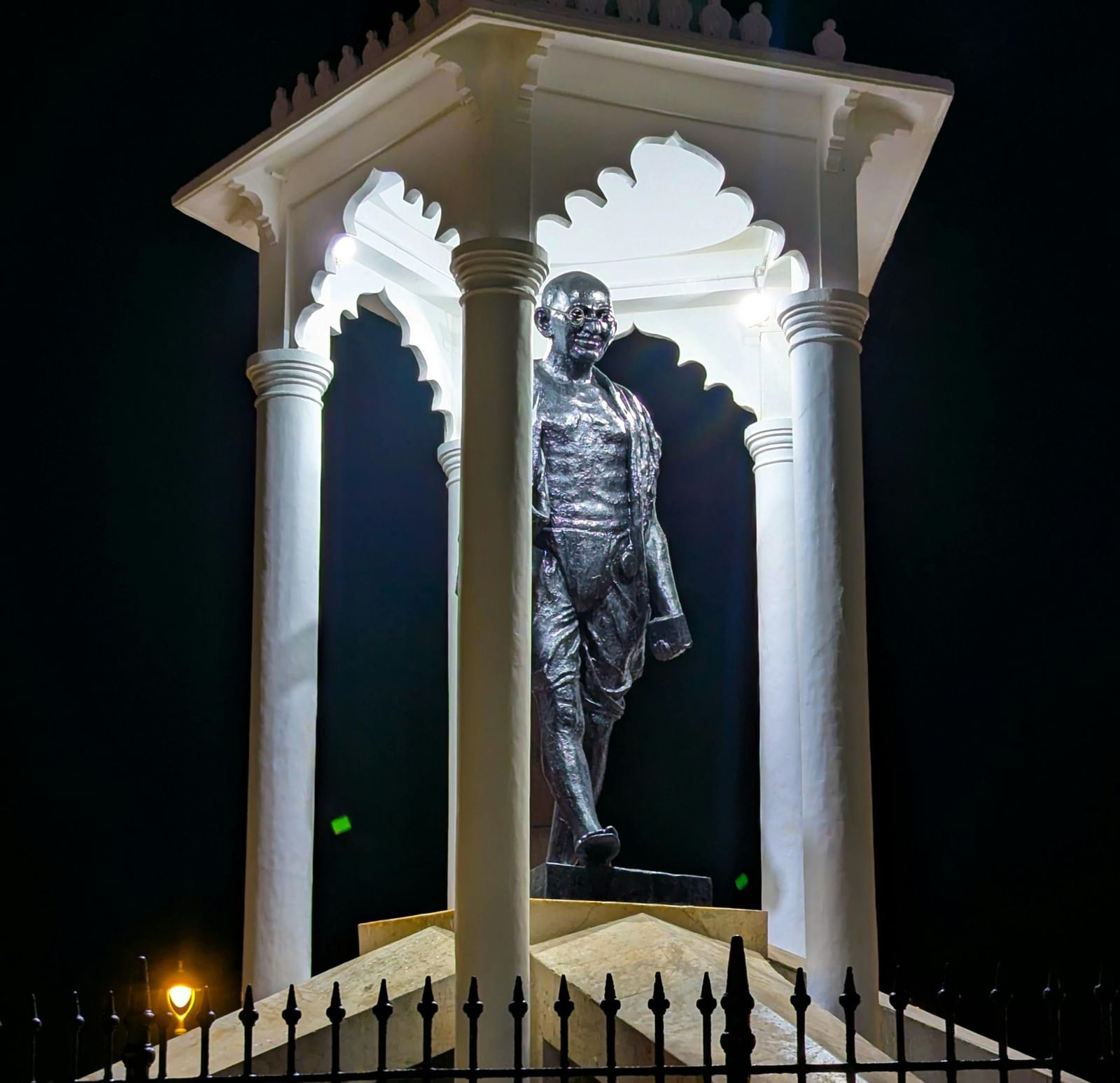 A tall white monument illuminated at night, showcasing an elegant statue beneath its glowing canopy.