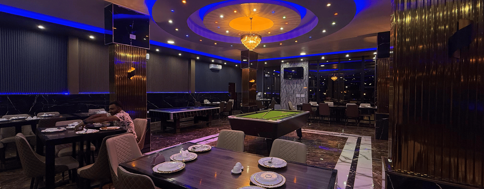 A dark, luxurious recreation room with a pool table, set tables, and illuminated by a central chandelier and blue neon ceiling lights at Luffu Club.