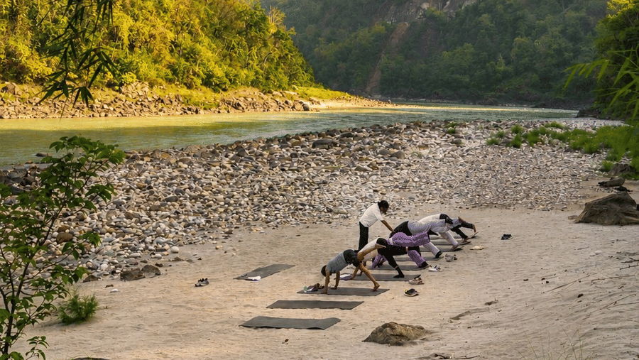 Guests practising yoga on mats along the sandy riverbank at Neemrana's Glasshouse on The Ganges - 21st Century, Rishikesh.