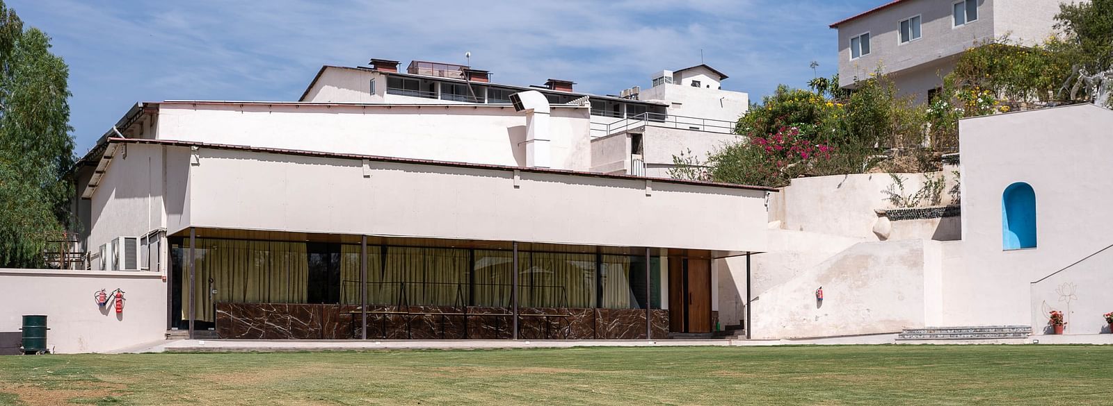 A facade of the white building of Bamboo Saa Resort & Spa, Udaipur with a flat roofline and a blue door set against a blue sky with white clouds.