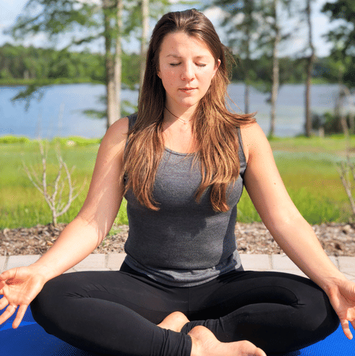 Woman meditating outdoors on a blue yoga mat in a cross-legged pose, with eyes closed and a serene lake view behind her