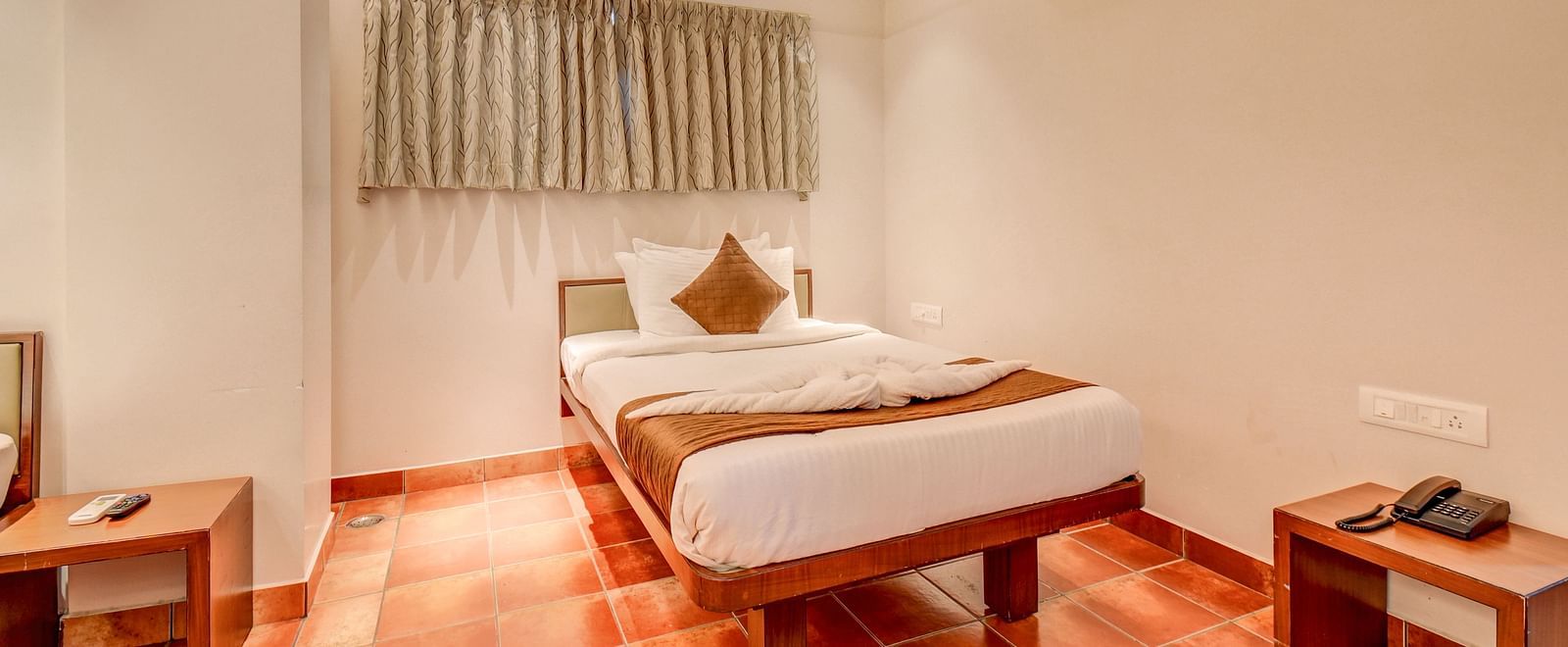 A spacious room at Harigovinda's JNStay with a bed, two wooden side tables, a wall-mounted AC, and high windows covered with curtains