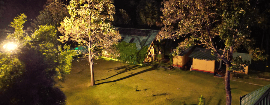 The illuminated facade of Ibex Resorts, Malampuzha (Kava Eco Camp and Caravan Park), featuring trees, and greenery at night.