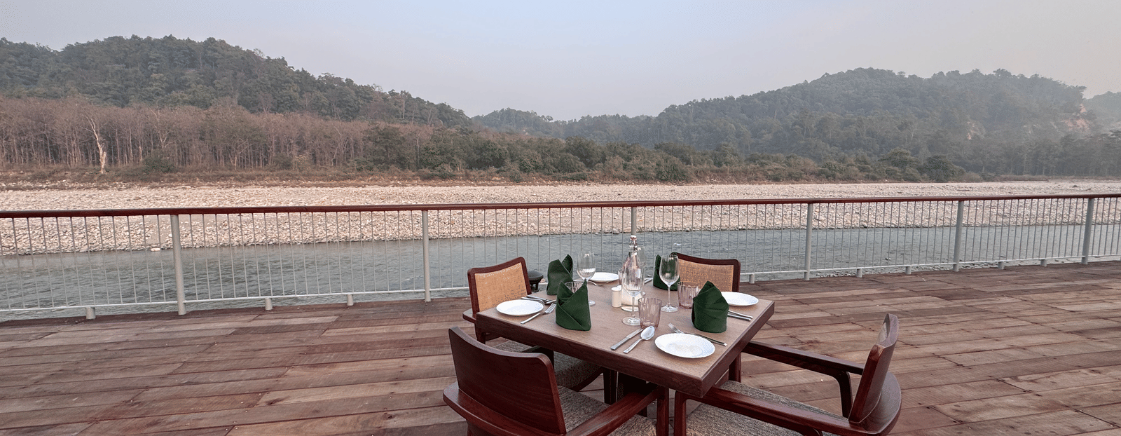 An elegant dining setup for four on a wooden deck overlooking a wide, flowing river and forested hills.