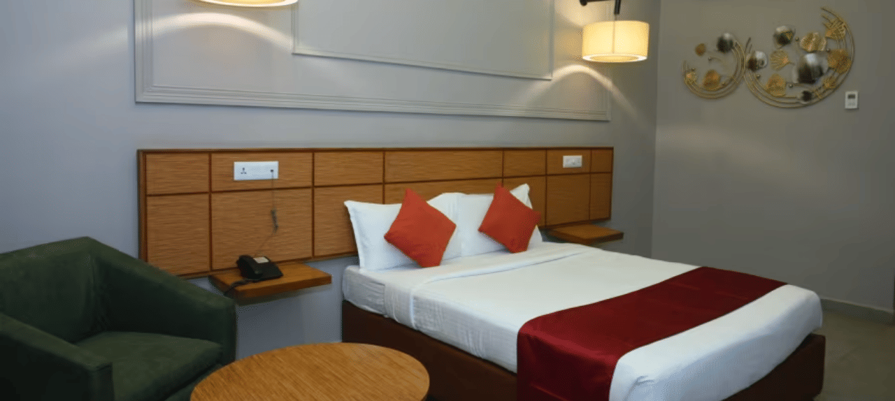 Bedroom with double bed, red runner, pillows, green chair, table and wall lamps at Max Hotel Prayagraj.