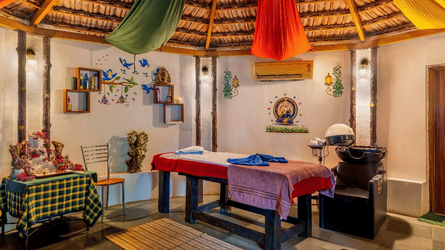 A spa room featuring a thatched roof with colourful draped fabrics, a massage table with blue towels, and shelving at Amantra Shilpi Resort, Udaipur.