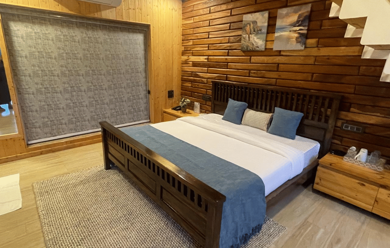 Megha Resort & Villas, Dehradun Standard Room, presenting a spacious interior with a prominent timber accent wall, a large bed with a fabric throw, and a textured area rug on a light wooden floor.