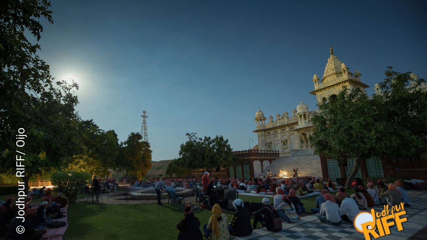 View of Jodhpur with a monument illuminated under the full moon, visitors exploring the site, and some seated at night, enjoying the serene atmosphere.