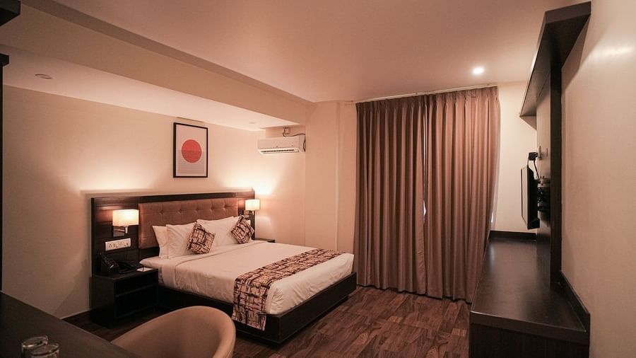 Queen size bed in the Deluxe Room of VITS Select, Bengaluru, also featuring a wall-mounted television, wooden flooring, a work desk, air-conditioning, and closed curtains.