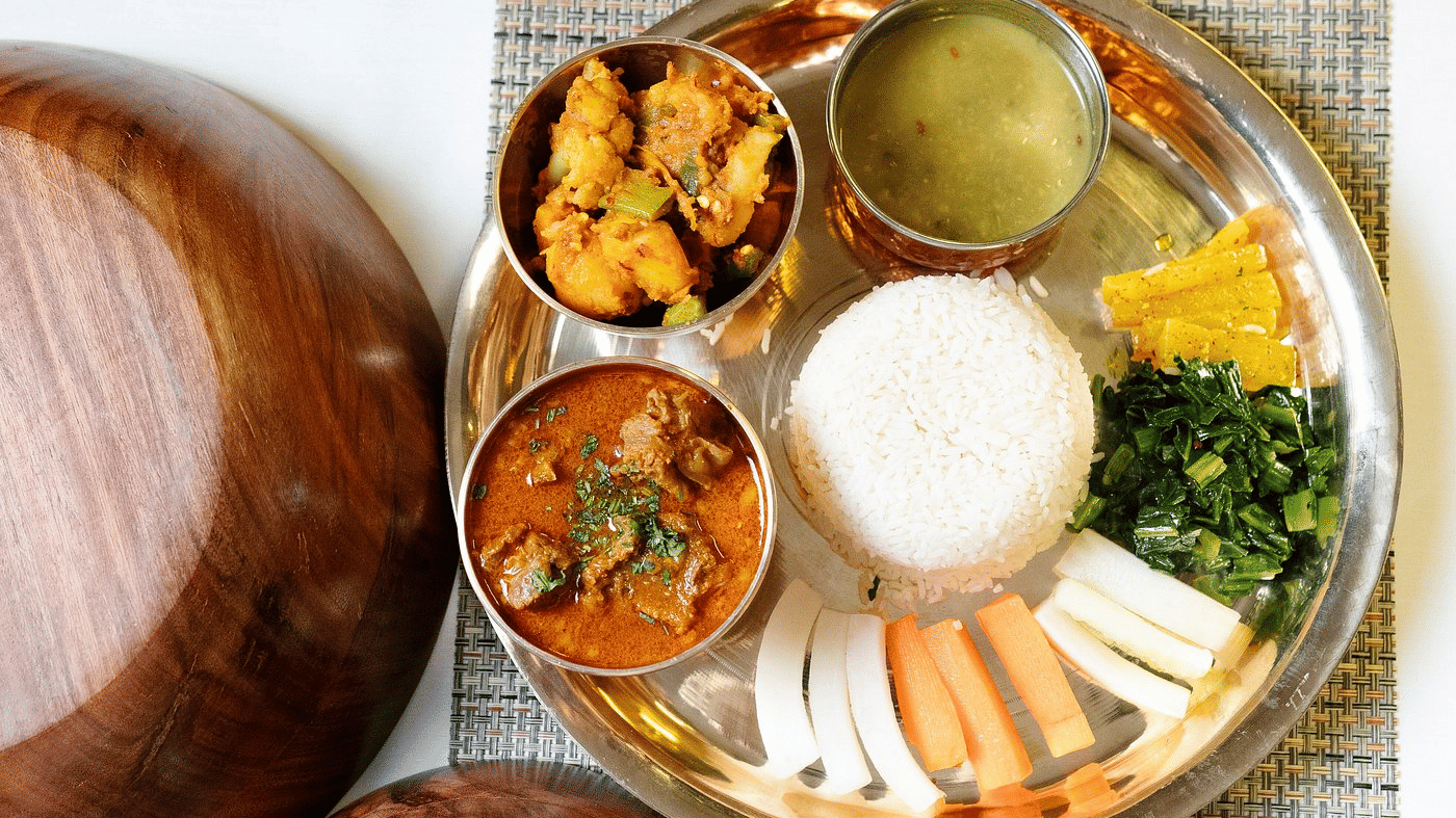 Overhead Nepali thali with rice, mutton curry, spiced potatoes, greens, radish, carrot and lentil soup on a brass platter at Ila Comfort, Baglung.