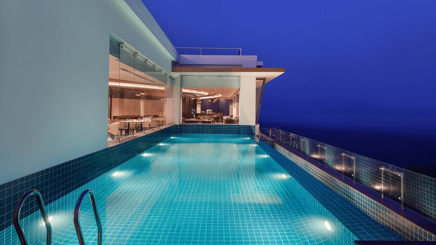 Outdoor tiled pool at dusk with underwater lighting, a glass safety fence and the lit restaurant alongside at Ila Resort, Maulakali.