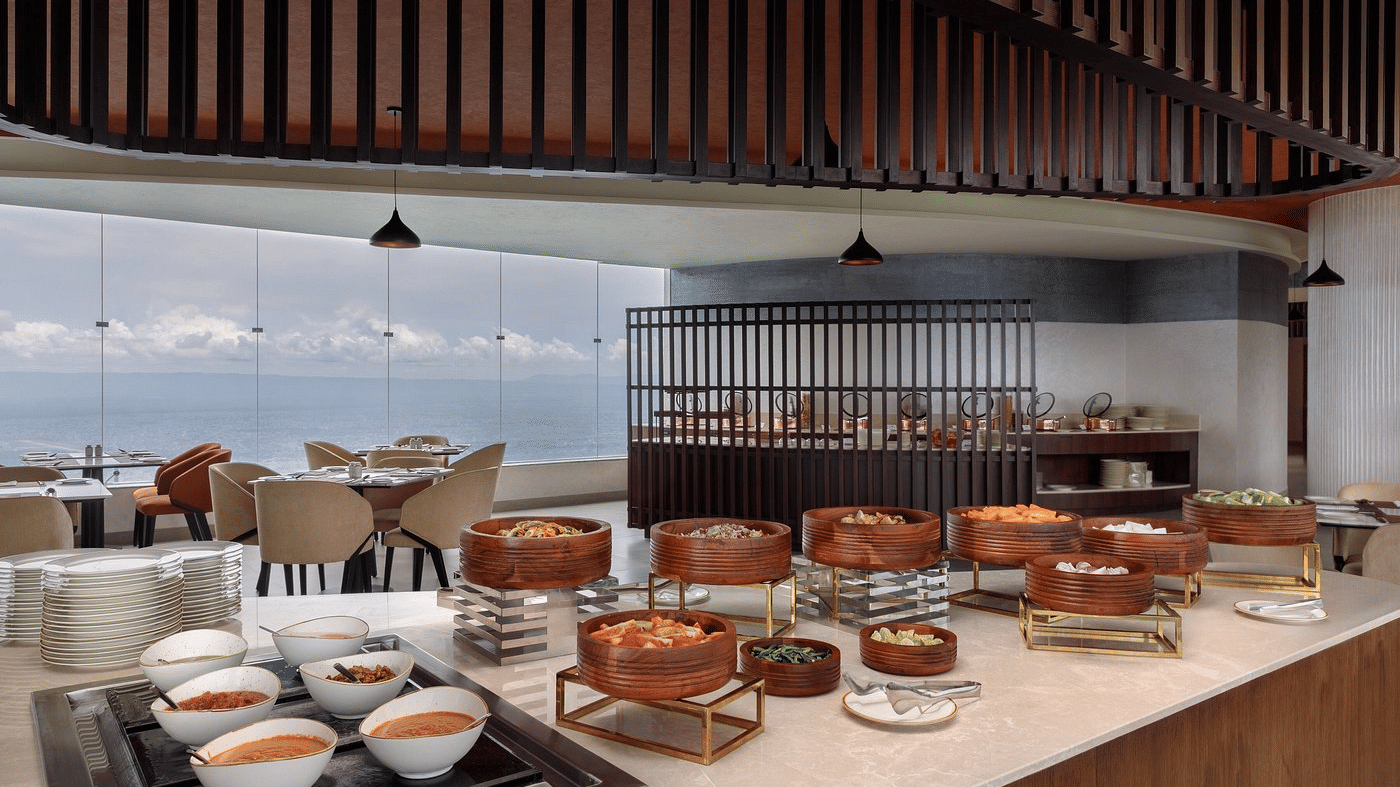 Restaurant buffet counter with wooden serving bowls on stands and dining tables beside floor-to-ceiling windows at Ila Resort, Maulakali.