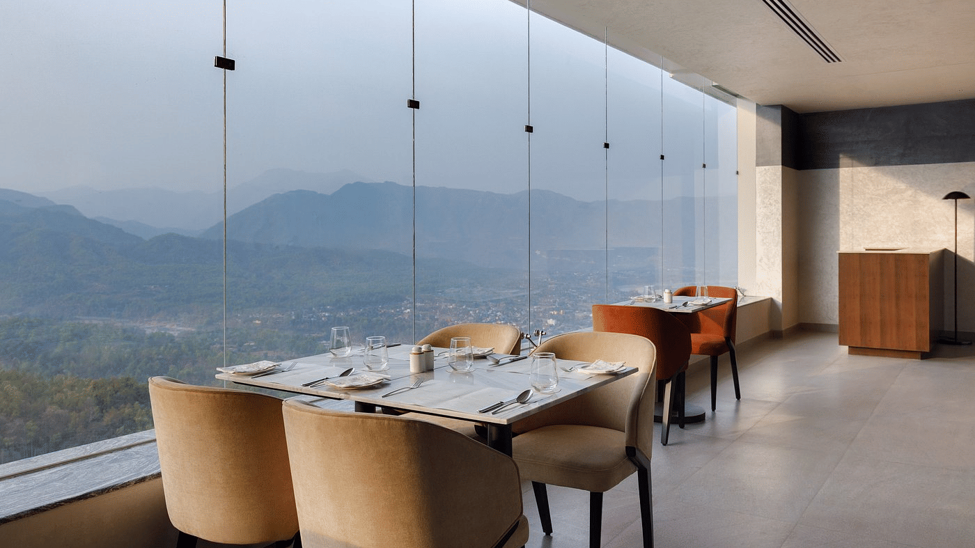 Restaurant tables laid with cutlery beside floor-to-ceiling glazing, with views over the valley town and hills, at Ila Resort, Maulakali.