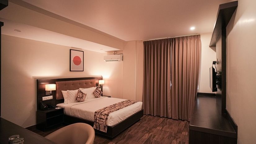 Queen size bed in the Deluxe Room of VITS Select, Bengaluru, also featuring a wall-mounted television, wooden flooring, a work desk, air-conditioning, and closed curtains.