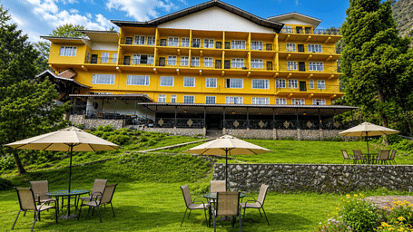The multi-storey facade of Orsino Khangri Karpo Retreat & Spa, Lachung during the day featuring a lawn with sets of tables and umbrellas for guests.