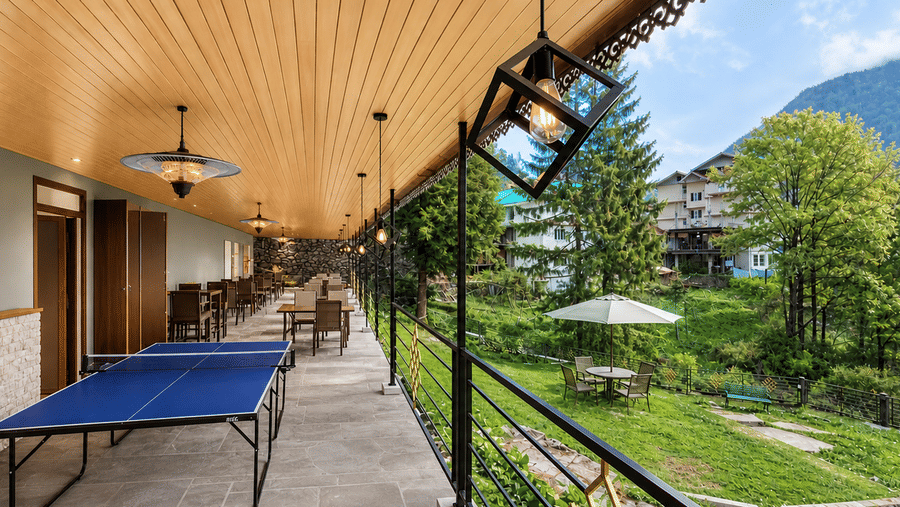 Opal bar at Orsino Khangri Karpo Retreat & Spa, Lachung featuring a long outdoor walkway with a table tennis table and several seating areas. A railing overlooks a garden and forested hills.