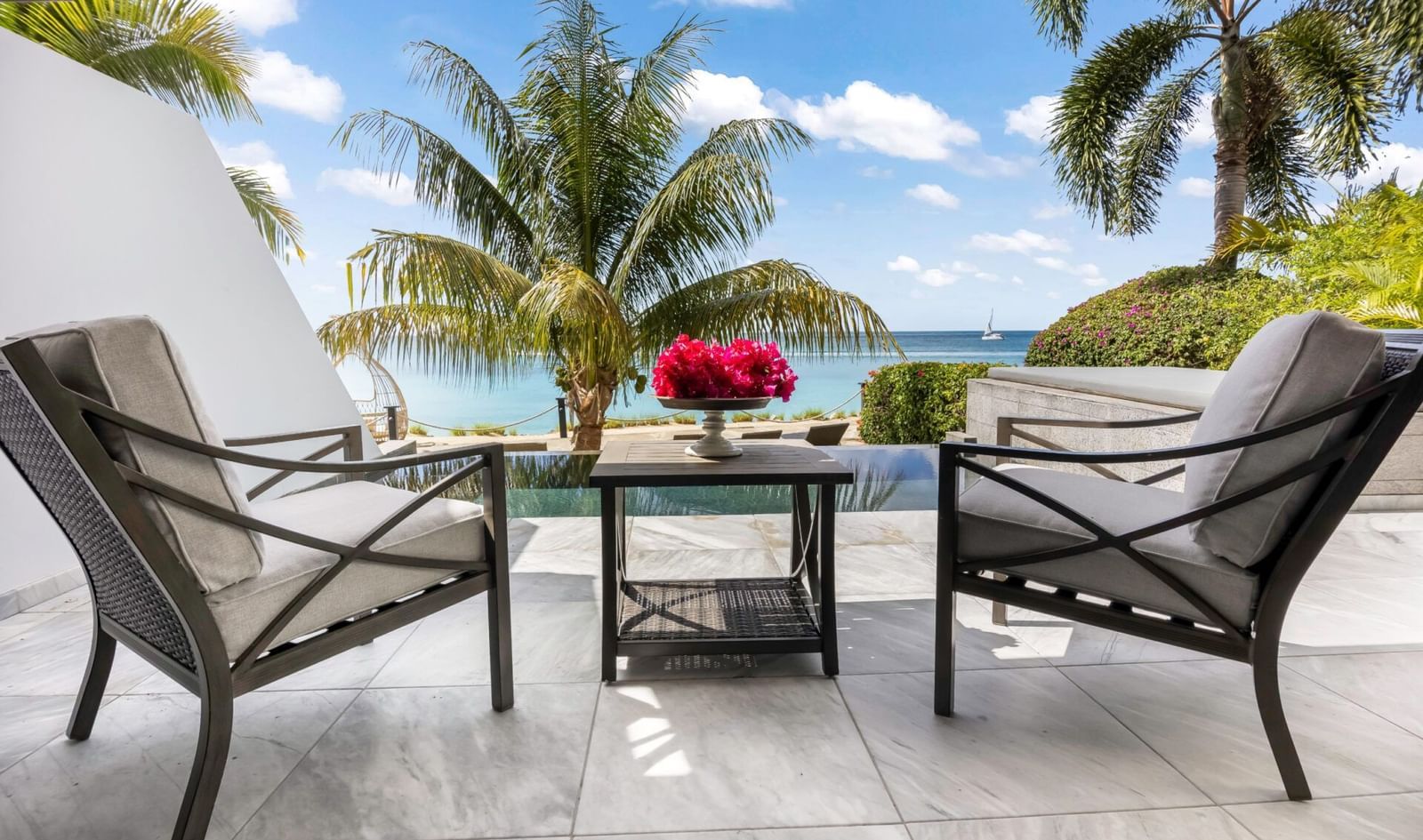 Two lounge chairs facing a pool featuring coconut trees and blue sky in its backdrop.