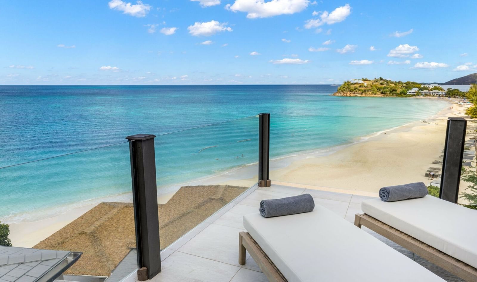 A stunning view from a modern balcony with glass railings, overlooking a pristine white sand beach, turquoise ocean, and distant coastline under a blue sky. Two lounge chairs with rolled towels are on the tiled balcony.