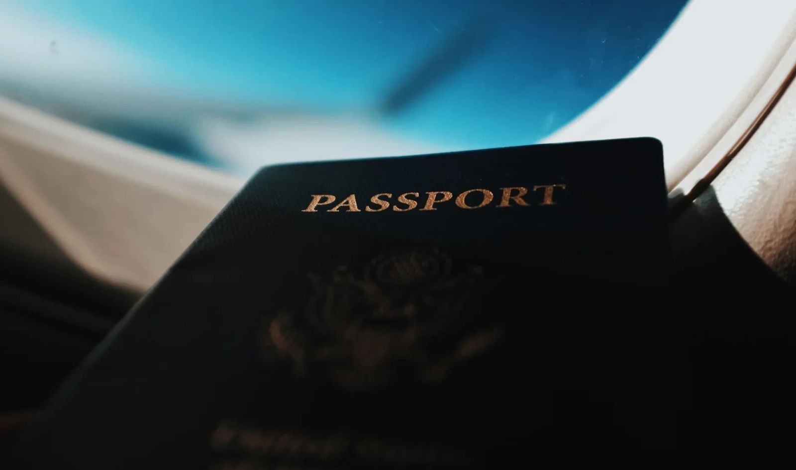 A passport rests near an airplane window, with the blurred wing and blue sky visible outside.