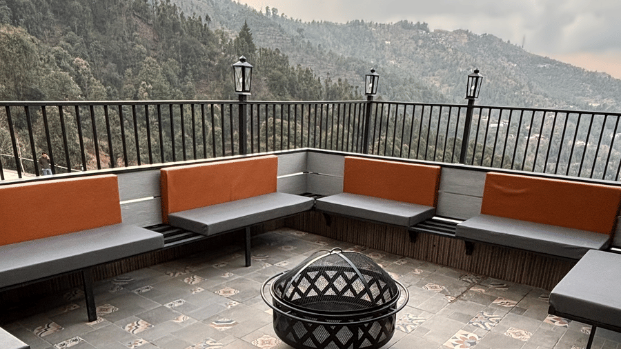 Terrace fire pit under a mesh cover, ringed by L-shaped benches with grey and orange cushions, railings and lamp posts, at The Chamomile Estate.