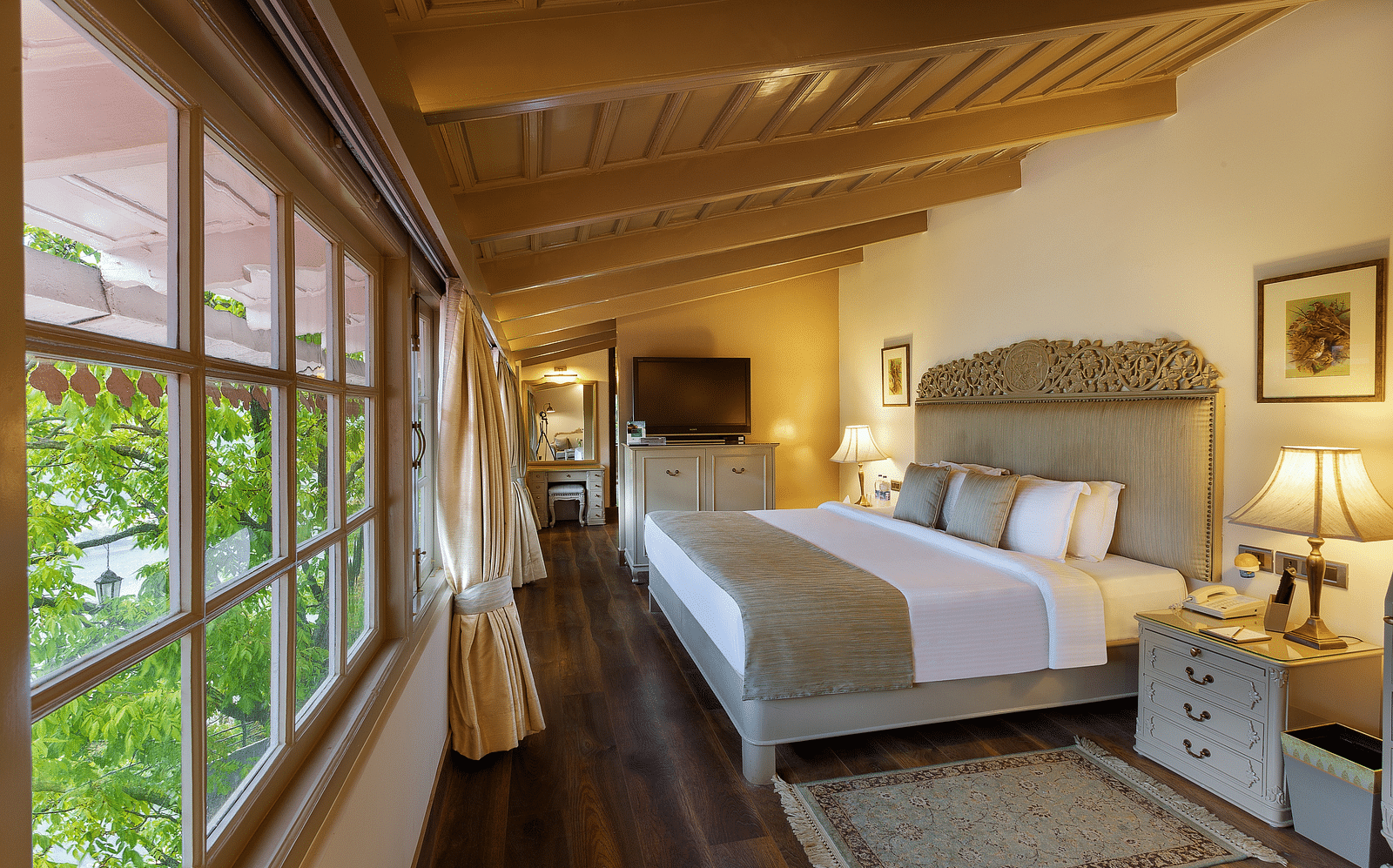 Image of a cosy room with lush decors, king size bed, wooden ceiling and  windows on the side and framed pictures on the other side- The Naini Retreat