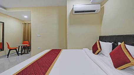 A hotel bedroom with double bed featuring white linens and red accent pillows, a black upholstered headboard, a mirror, two orange study chairs and a simple, uncluttered decor.