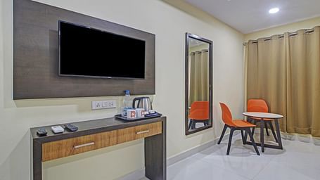 A neat hotel room with a wall-mounted TV, a wooden entertainment console, and a casual seating area with a vibrant orange chair.