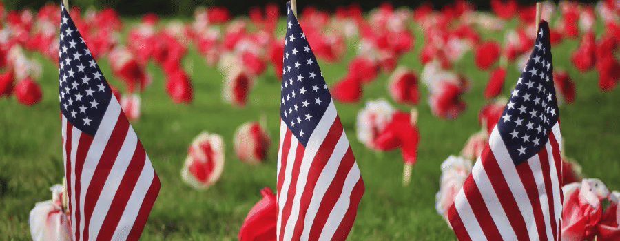 Three small American flags planted in a grassy field with a backdrop of blurred red and white flowers
