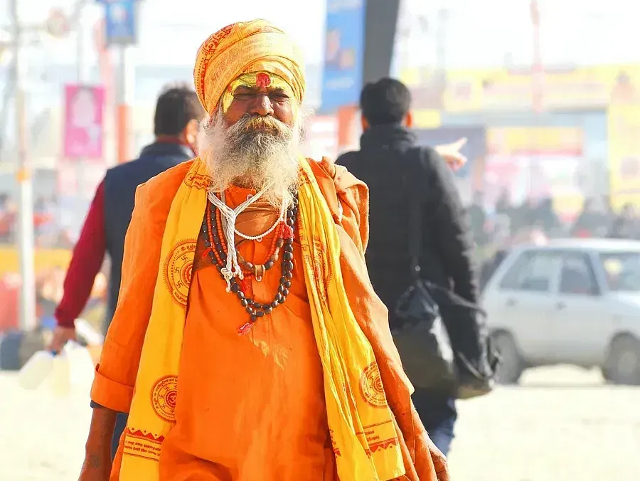 An elderly Hindu ascetic (sadhu) wearing orange robes and beads, walking through a busy street.