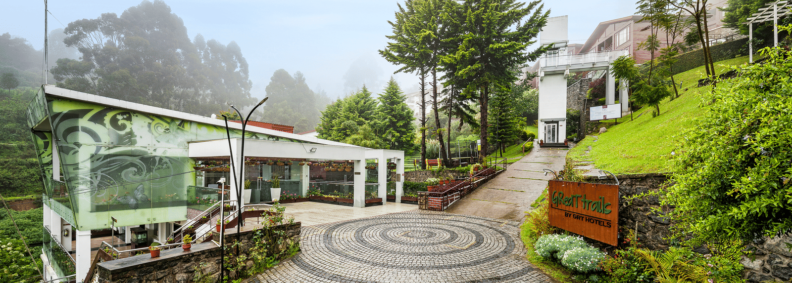 A facade image of the GReaT Trails Kodaikanal By GRT Hotels with lush greenery and the trails leading to the best mountain view resort, Kodaikanal, Tamil Nadu