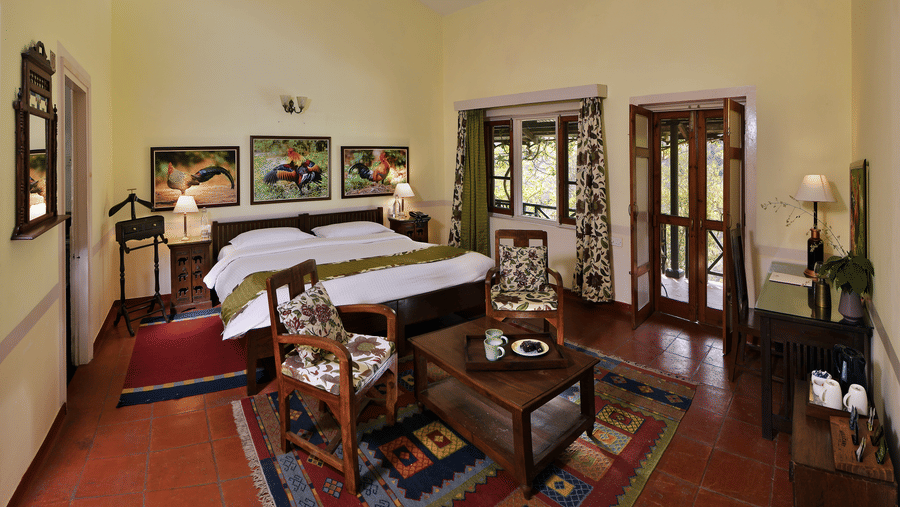 Spacious heritage style suite with a large bed wooden furniture seating area patterned rugs tall ceiling fans and natural light creating a calm and comfortable stay experience at Neemrana’s Glasshouse on The Ganges surrounded by a peaceful indoor atmospher