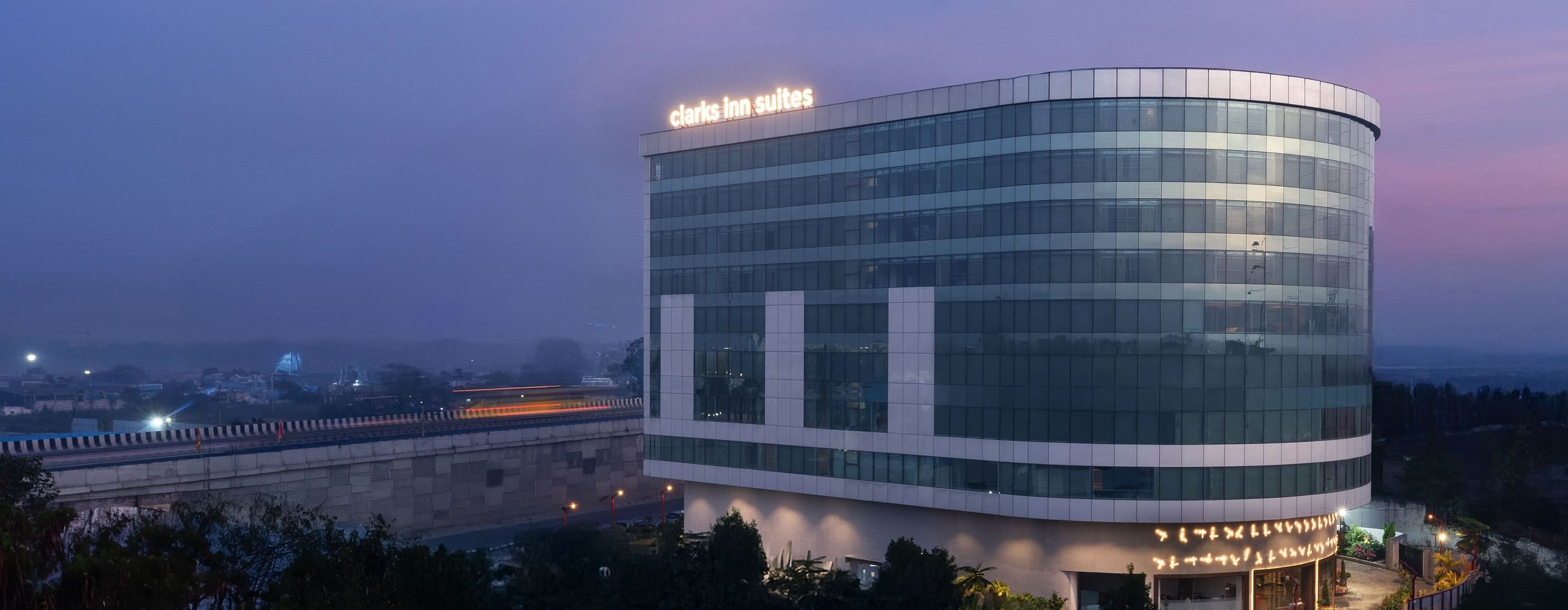 Evening shot of a curved glass hotel building with illuminated signage against a dusky sky at Clarks Inn Suites, Hyderabad