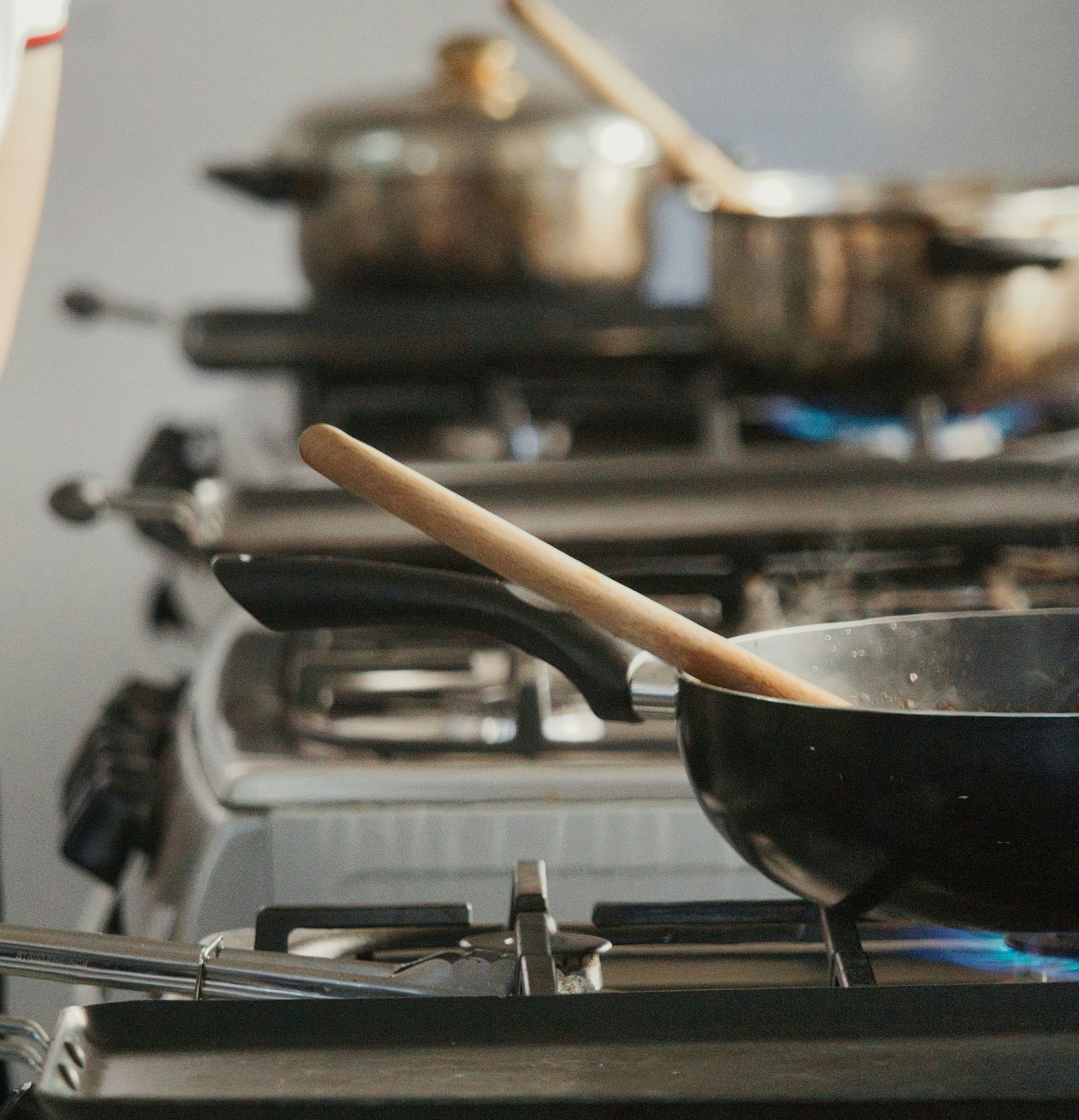 A close-up of gas stoves with blue flames heating several pots and pans, including a wok with a wooden spatula, in a kitchen setting during a cooking class or meal preparation.