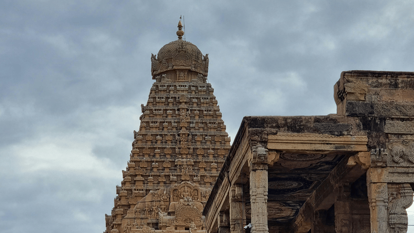 Tourists and pilgrims gather at the ancient Brihadeeswarar Temple, admiring its grand Dravidian architecture under a cloudy sky. It is one of the best places to see around Thanjavur.
