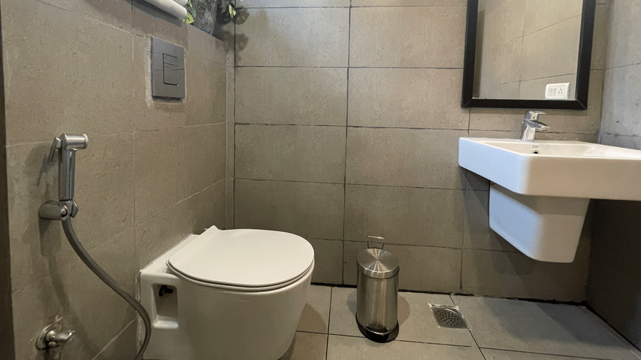 A bathroom at Sanctum Suites, Richmond Road, Bangalore has a toilet, a white washbasin, a mirror, and a wall mounted rack with towel.