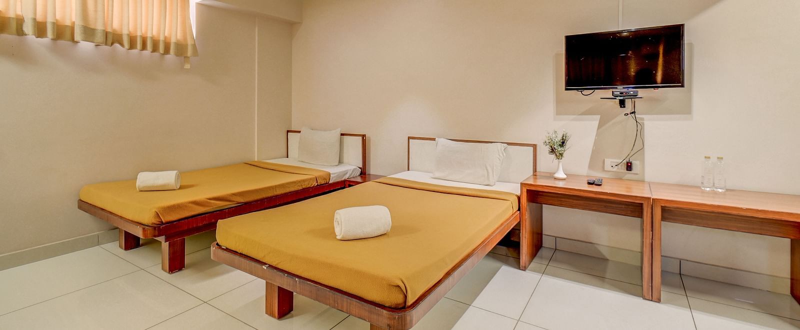 A spacious room at Harigovinda's JNStay, featuring a wall-mounted TV above a wooden table set against the wall, and two beds