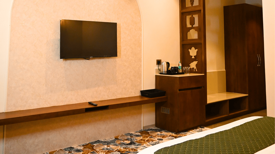 A wooden media console and wall-mounted TV in a Kadamb Kuteer room, with a glimpse of the bed and patterned floral carpeting.