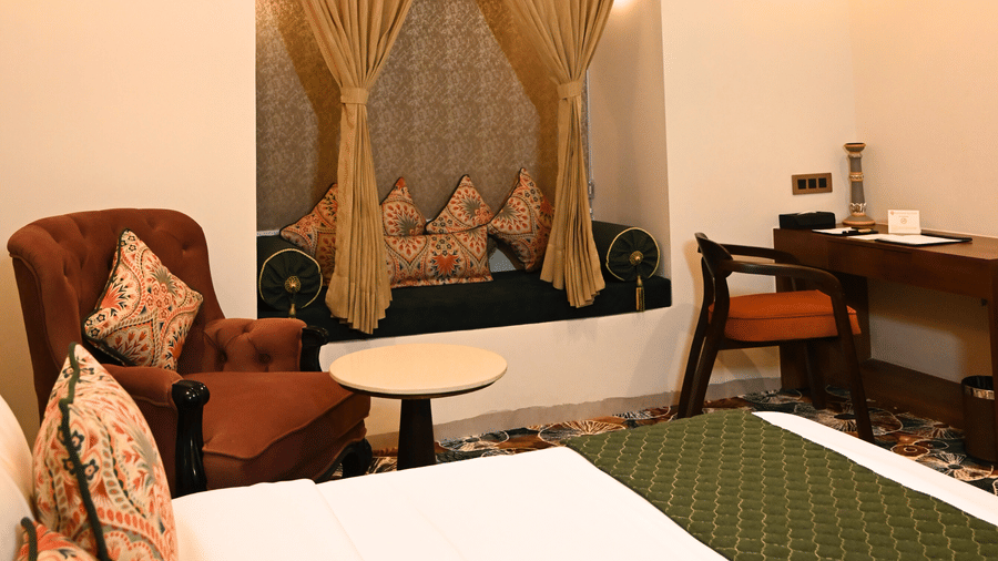A corner of a room at Kadamb Kuteer featuring a wooden chair, a small side table, and a framed wall hanging against a neutral-colored wall.